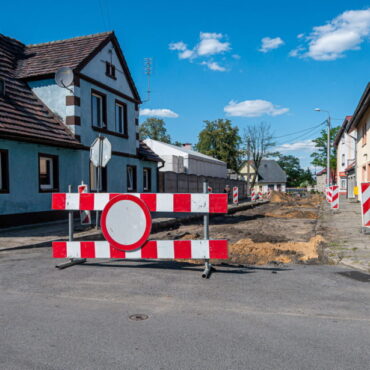 Rozkopana ulica, zagrodzona biało-czerwonymi barierkami i znakiem zakaz ruchu. Po obu stronach ulicy domy